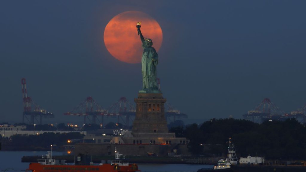 Full Harvest Moon setting behind the Statue of Liberty through a telephoto lens