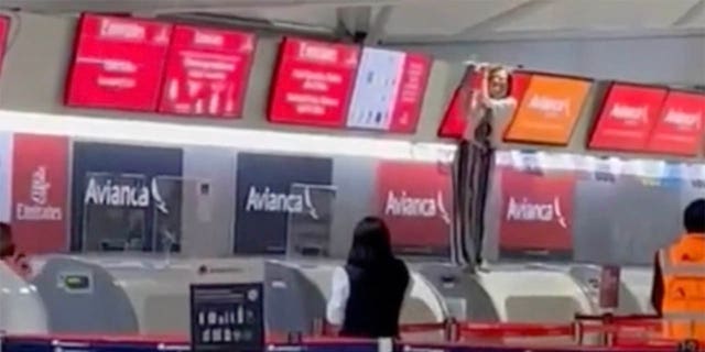 A repülőtér többi utasa messziről láthatja a kontrollon kívüli személyt - aki a check-in pultnál áll, és képernyőt tart fölötte.