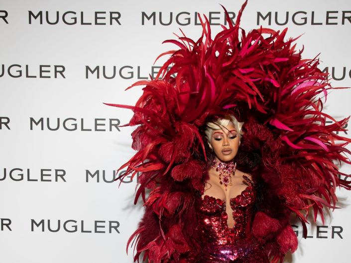 Cardi B rapper piros ruhában, piros tollakkal és piros köpennyel a fal előtt, amelyen az áll, hogy "  MUGLER "